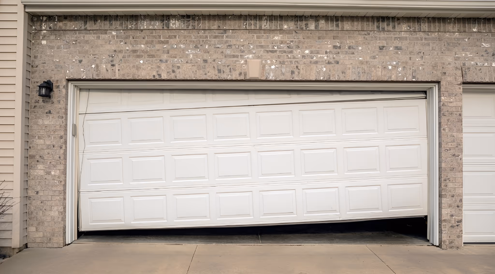 White garage door that is off-track and stuck at an angle, partially open, attached to a brick exterior of a residential home.
