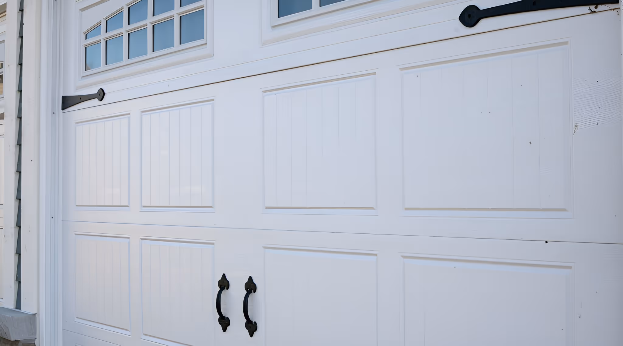 A white garage door with four rectangular panels and decorative black hardware on the bottom corners. The door features small windows at the top with a grid design.