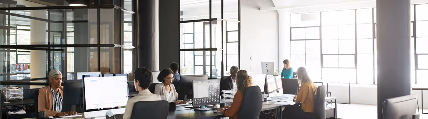 Escritório moderno com várias pessoas trabalhando em computadores em mesas compartilhadas ao lado de grandes janelas.