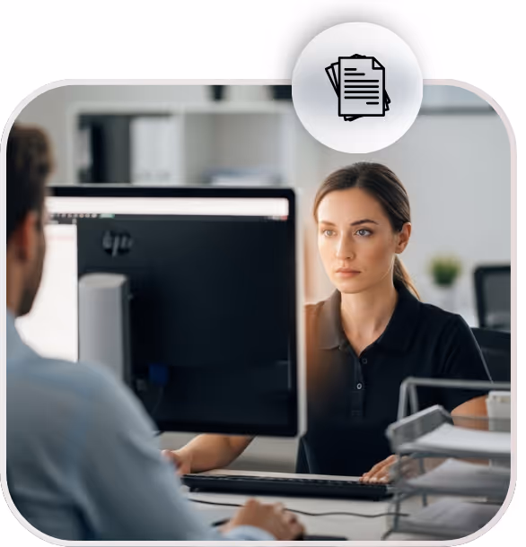 Mulher trabalhando em escritório olhando atentamente para a tela do computador, com um colega ao fundo.