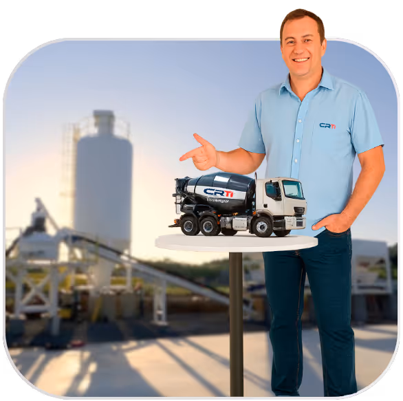 Homem sorridente em camisa azul clara apontando para um modelo em miniatura de caminhão betoneira sobre uma mesa, com uma planta de concreto desfocada ao fundo.