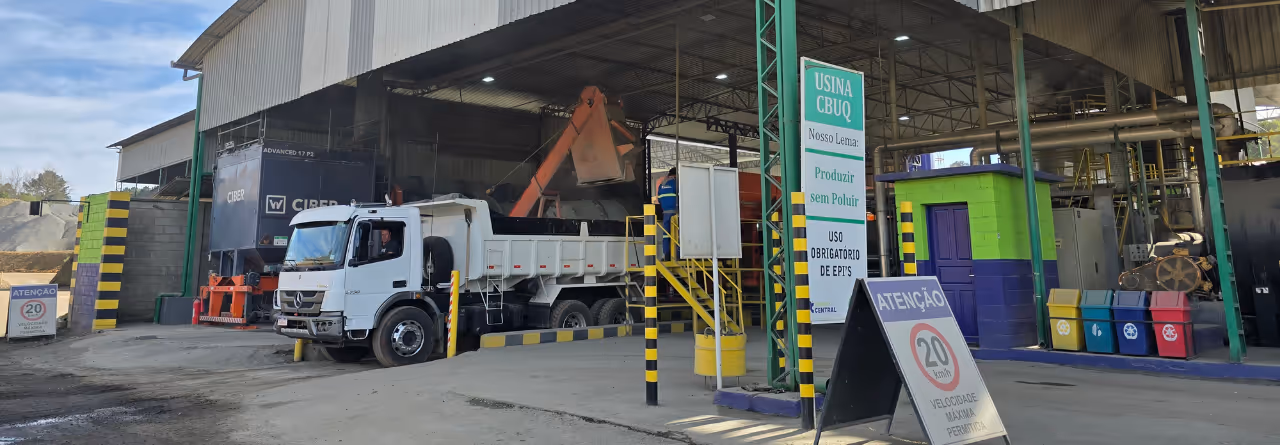 Caminhão branco dentro de uma usina de asfalto com máquina de carregamento e placas de segurança visíveis.