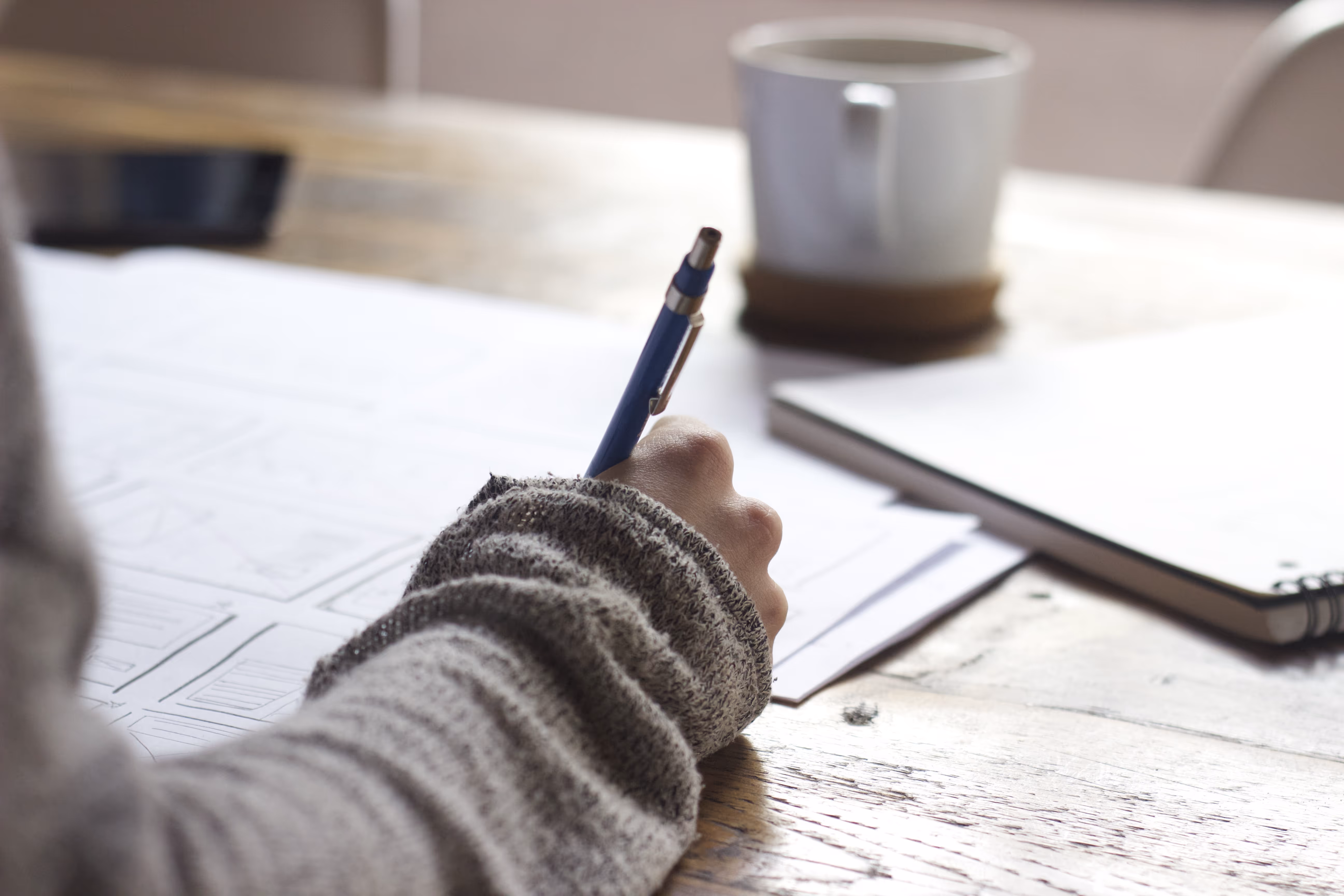 A womans hand on a wooden desk writing. A coffee cup is in the back of shot.