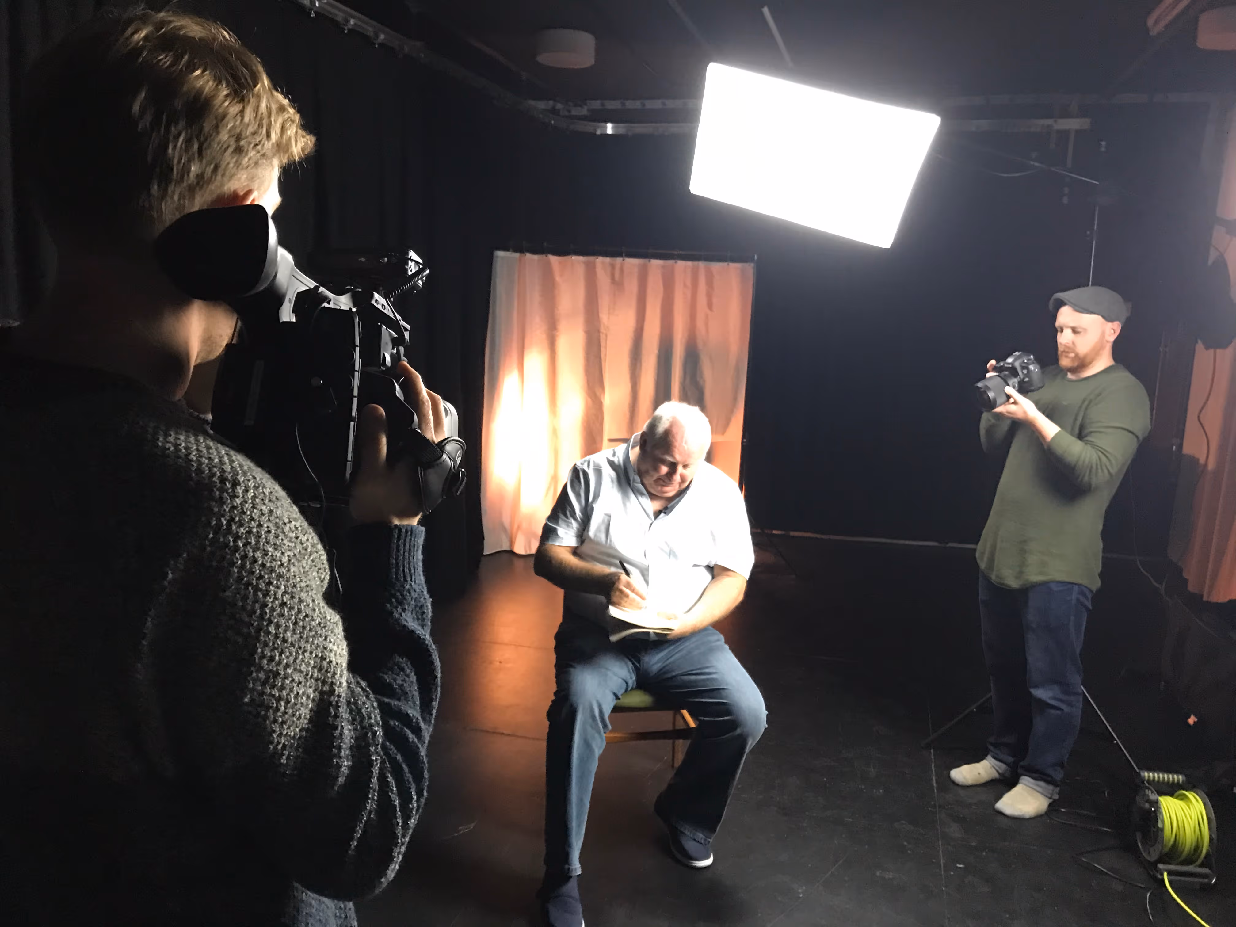 A photo of two cameramen filming a man who is sat in an interview drawaing something. Cameras, lights and tripods are in the shot.