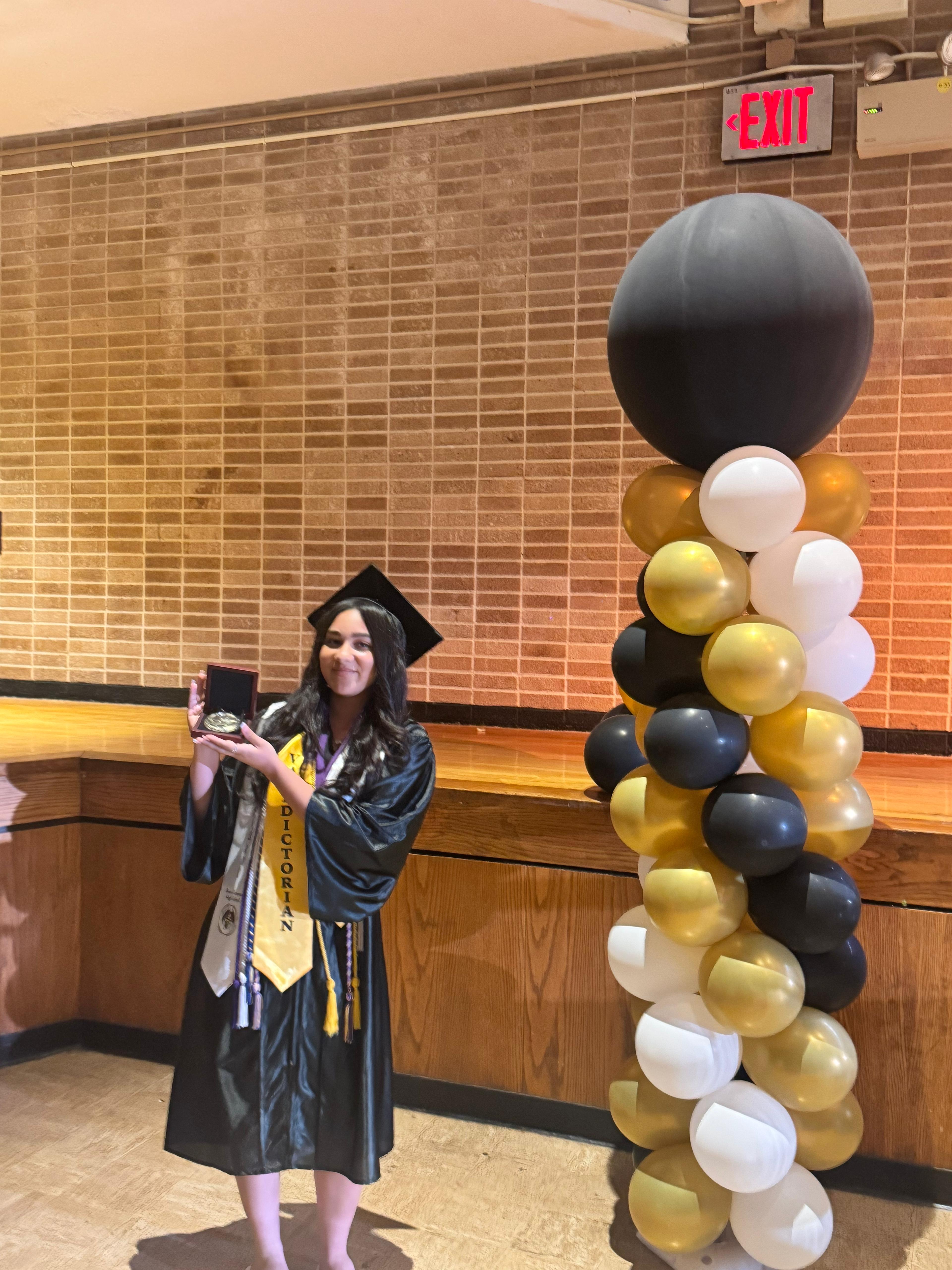 Valedictorian graduate holding award beside balloon column at ceremony