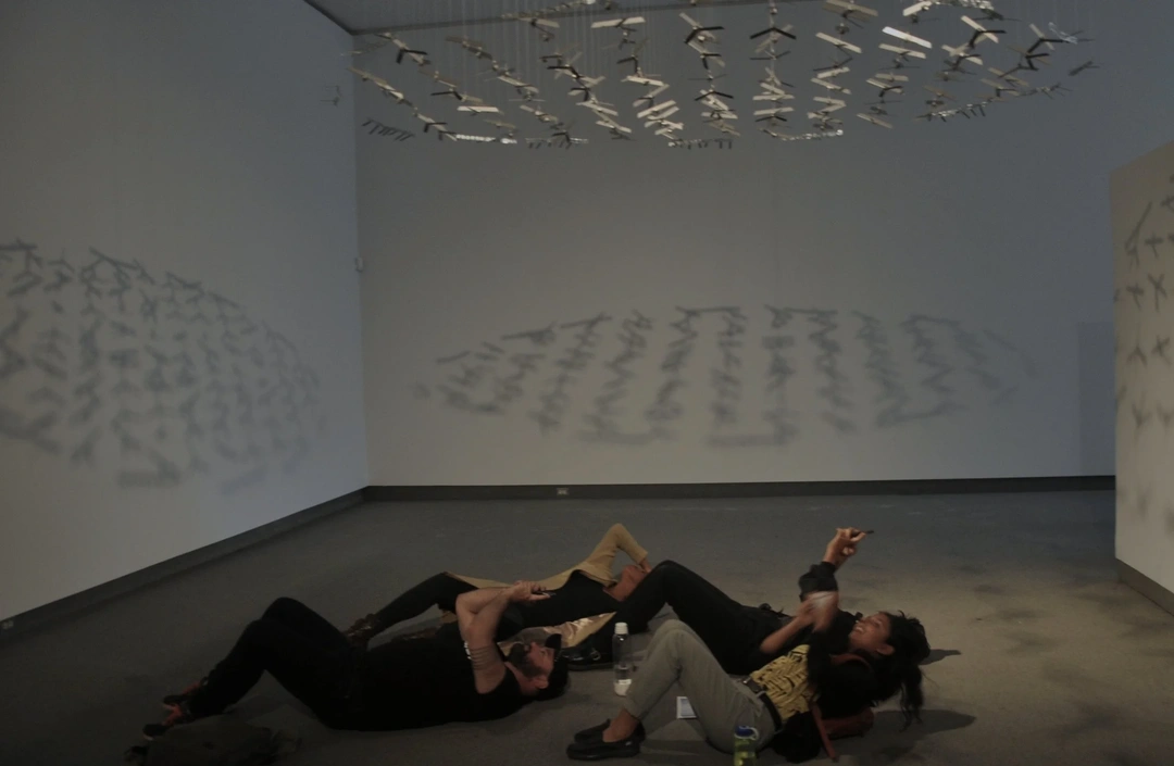 Installation view of Abdullah M. I. Syed's The Flying Rug with visitors lying on gallery floor beneath suspended currency carpets.