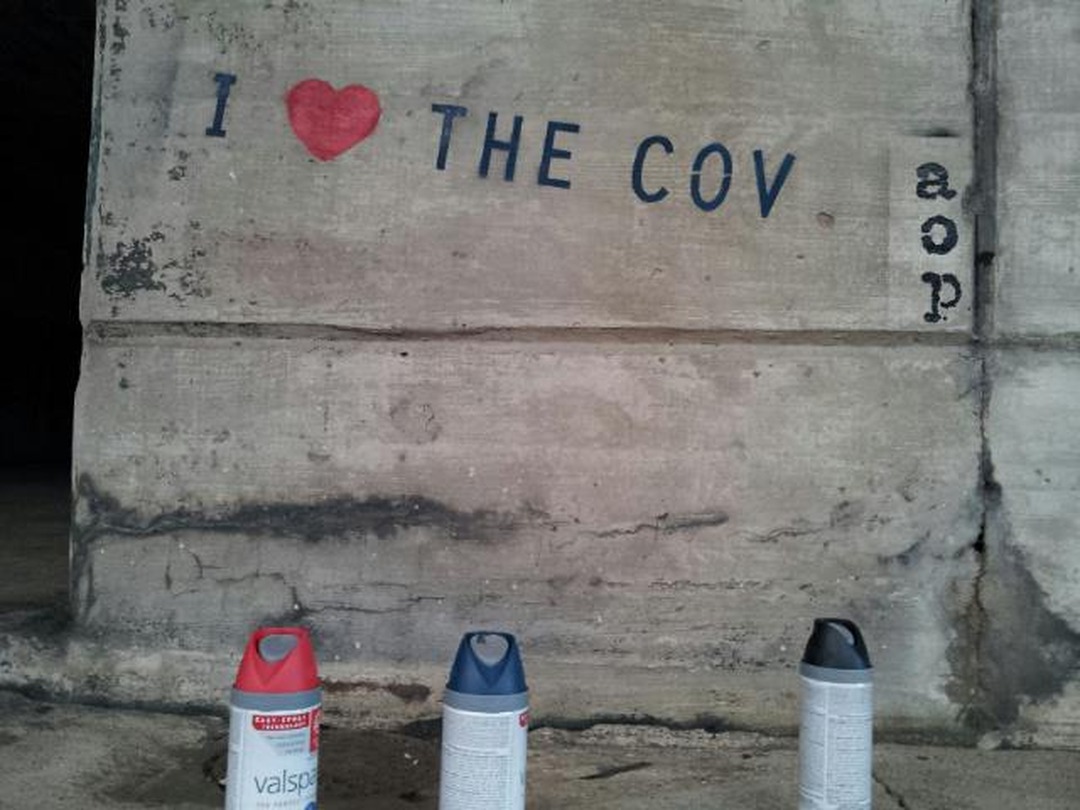 Spray-painted stencil on a concrete wall reading 'I love the Cov', with a small 'a o p' stamp and three spray cans below.