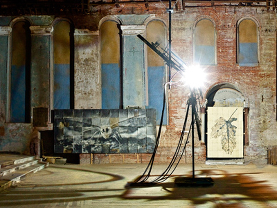 Interior of a brick church with tall arched windows, a bright carbon arc lamp in the center, and large photographic prints on the walls.