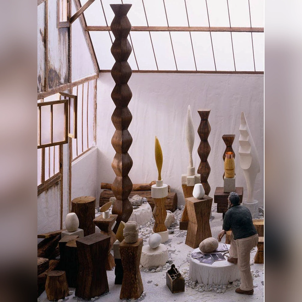 A miniature Brancusi stands with one foot on a stone slab among carved wooden columns and white and gold sculptures beneath a glass studio roof.