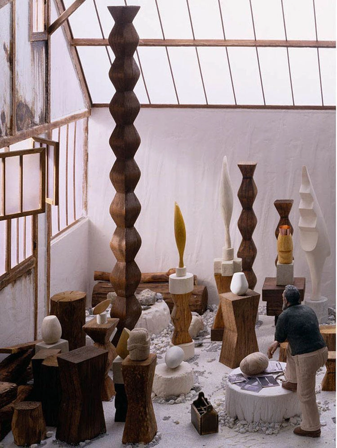 A miniature Brancusi stands with one foot on a stone slab among carved wooden columns and white and gold sculptures beneath a glass studio roof.