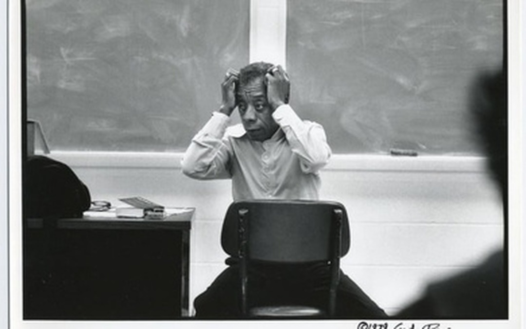 Black-and-white photograph of James Baldwin seated backwards on a chair, hands raised to his head.