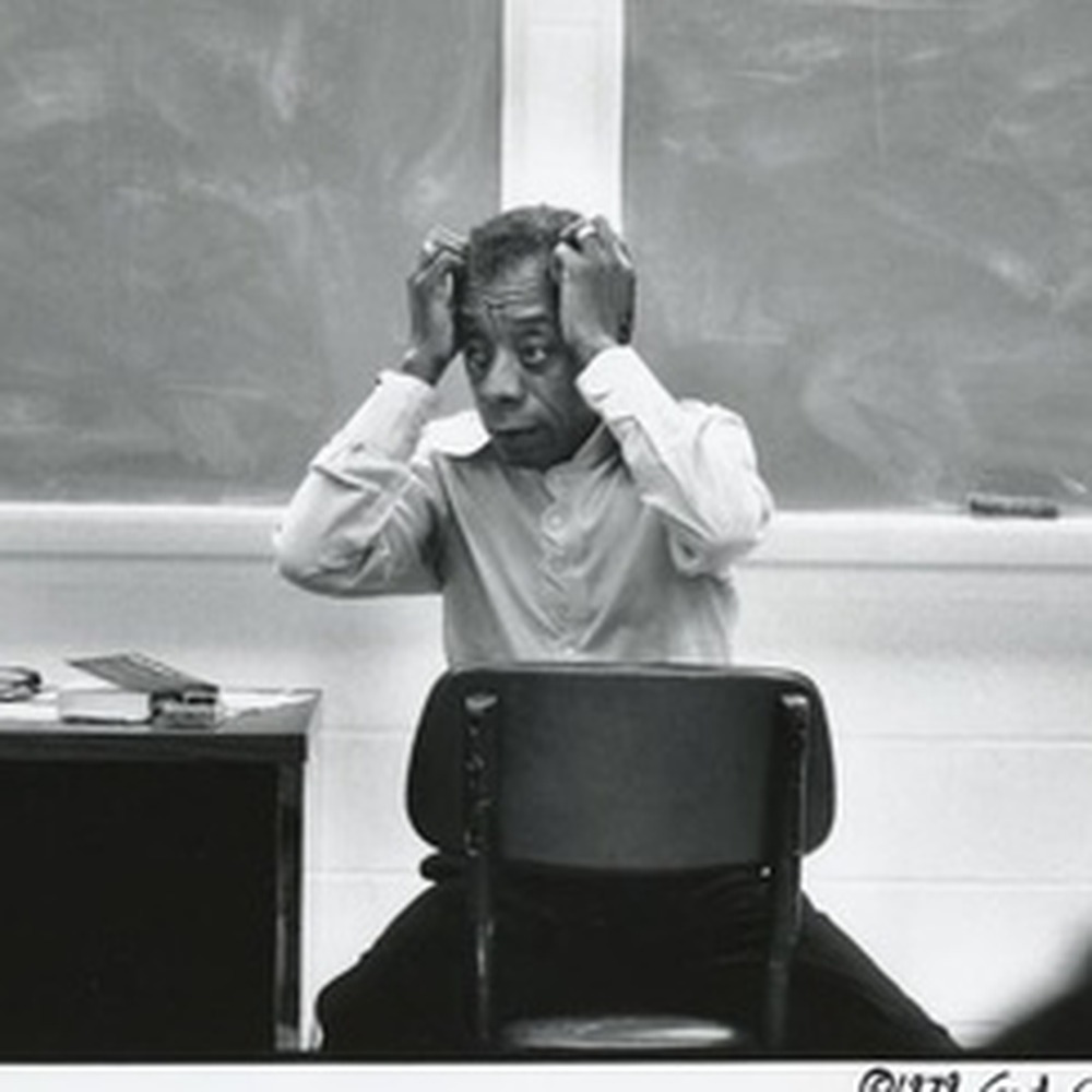 James Baldwin seated backwards on a chair, hands on his head.