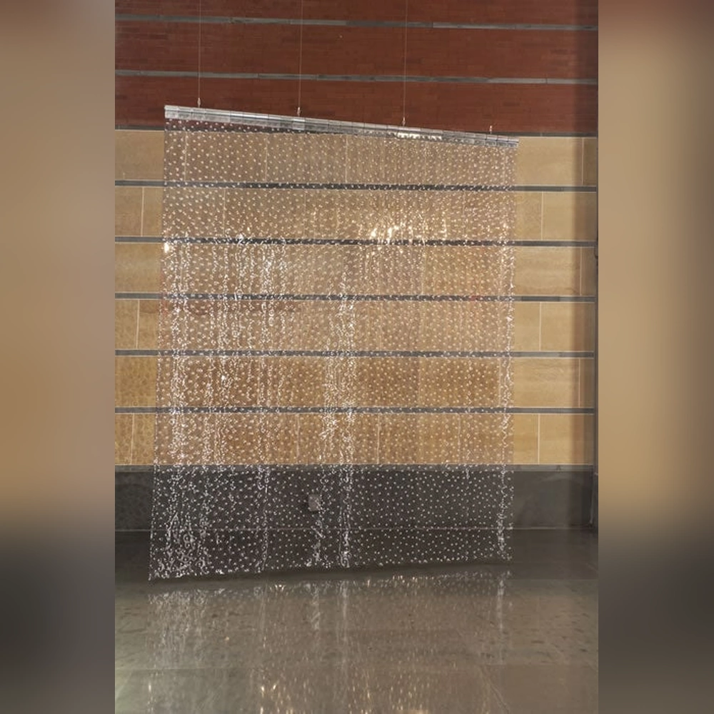 Translucent vertical PVC strips studded with silver rivets hang before a tan and brick gallery wall, reflected on the floor below.