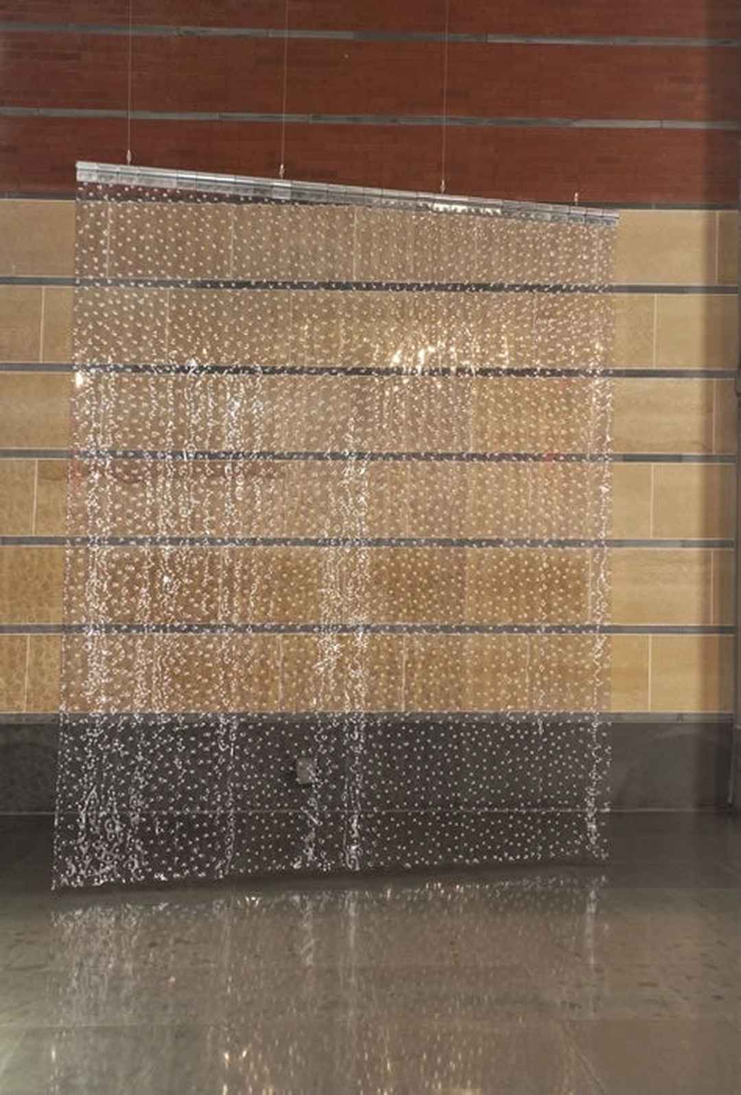 Translucent vertical PVC strips studded with silver rivets hang before a tan and brick gallery wall, reflected on the floor below.