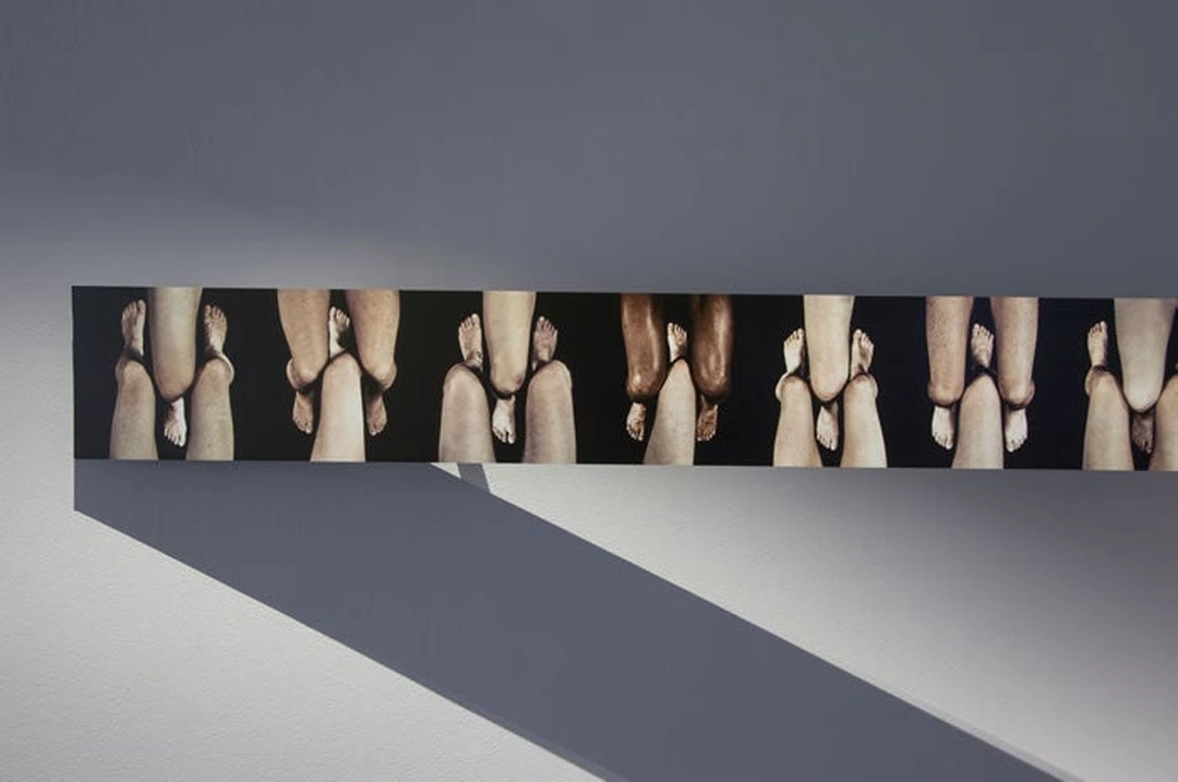 Detail of a horizontal photographic strip showing a sequence of touching knees on a gray gallery wall, with a diagonal shadow below. Photo by Tony Walsh, 2012. (Related image located during this archive's reconstruction.)