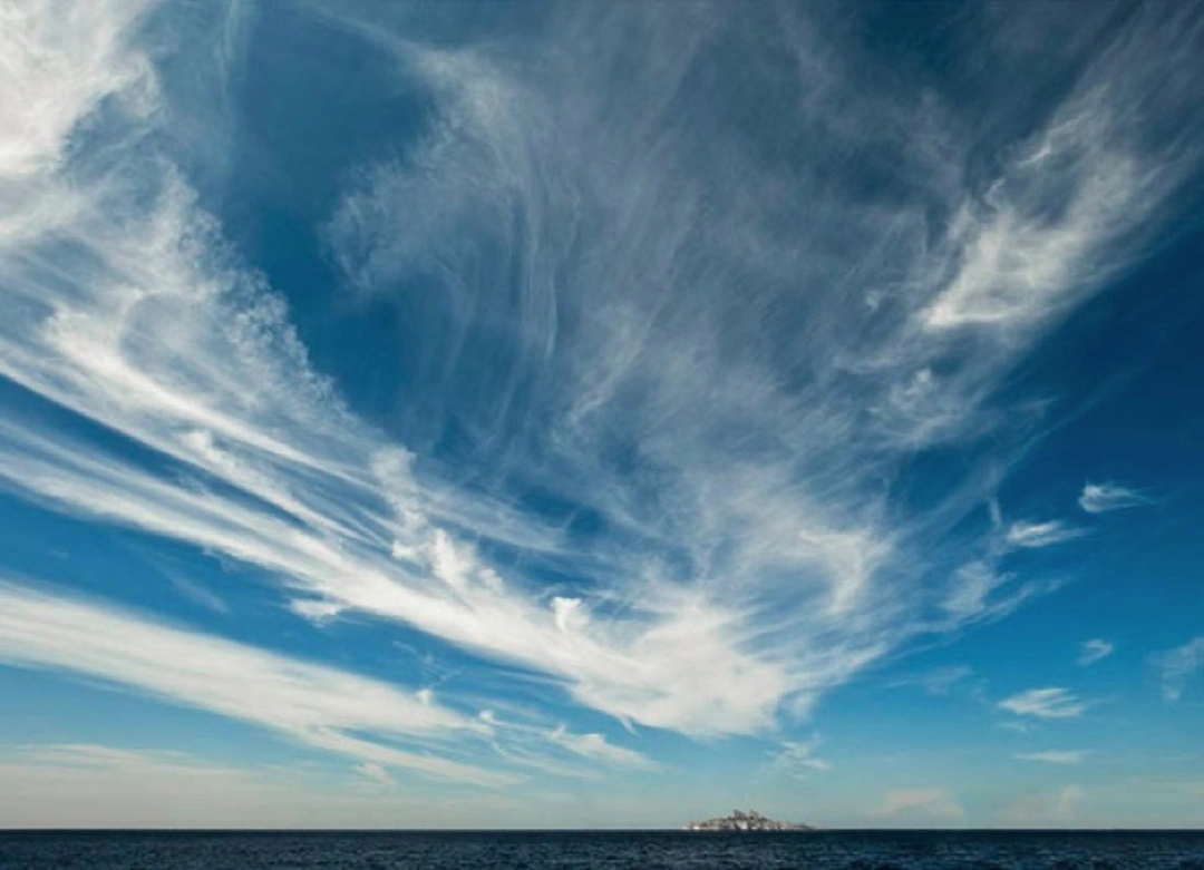 Wispy white clouds sweep across a turquoise sky above dark blue sea and a small distant rocky island near the horizon.