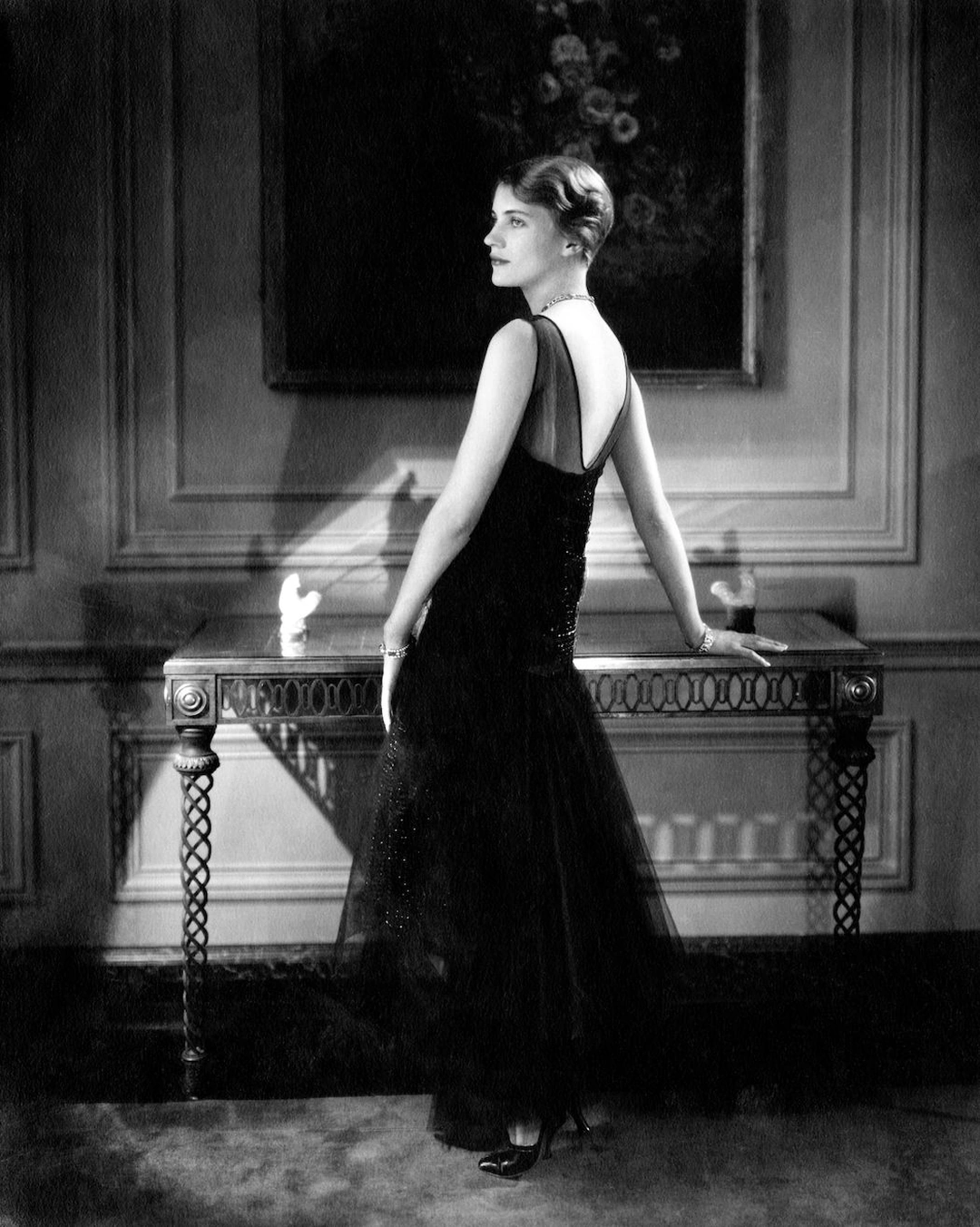 A woman in a long dark backless evening gown turns toward the camera with one hand resting on an ornate console table in a paneled room.
