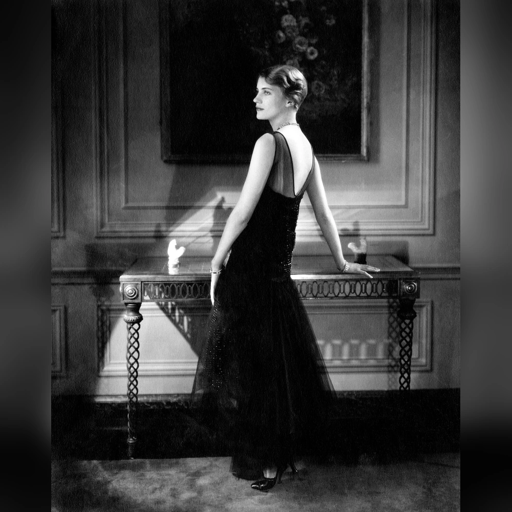 A woman in a long dark backless evening gown turns toward the camera with one hand resting on an ornate console table in a paneled room.