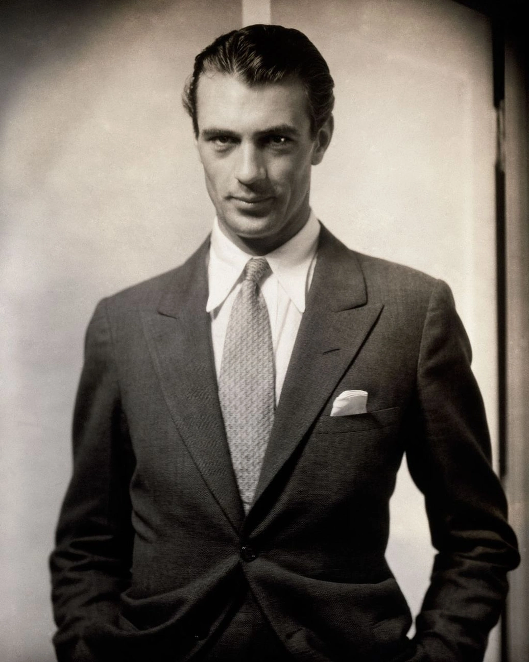A man in a dark suit, patterned tie and white pocket square stands with his hands in his pockets. Steichen “Gary-Cooper”