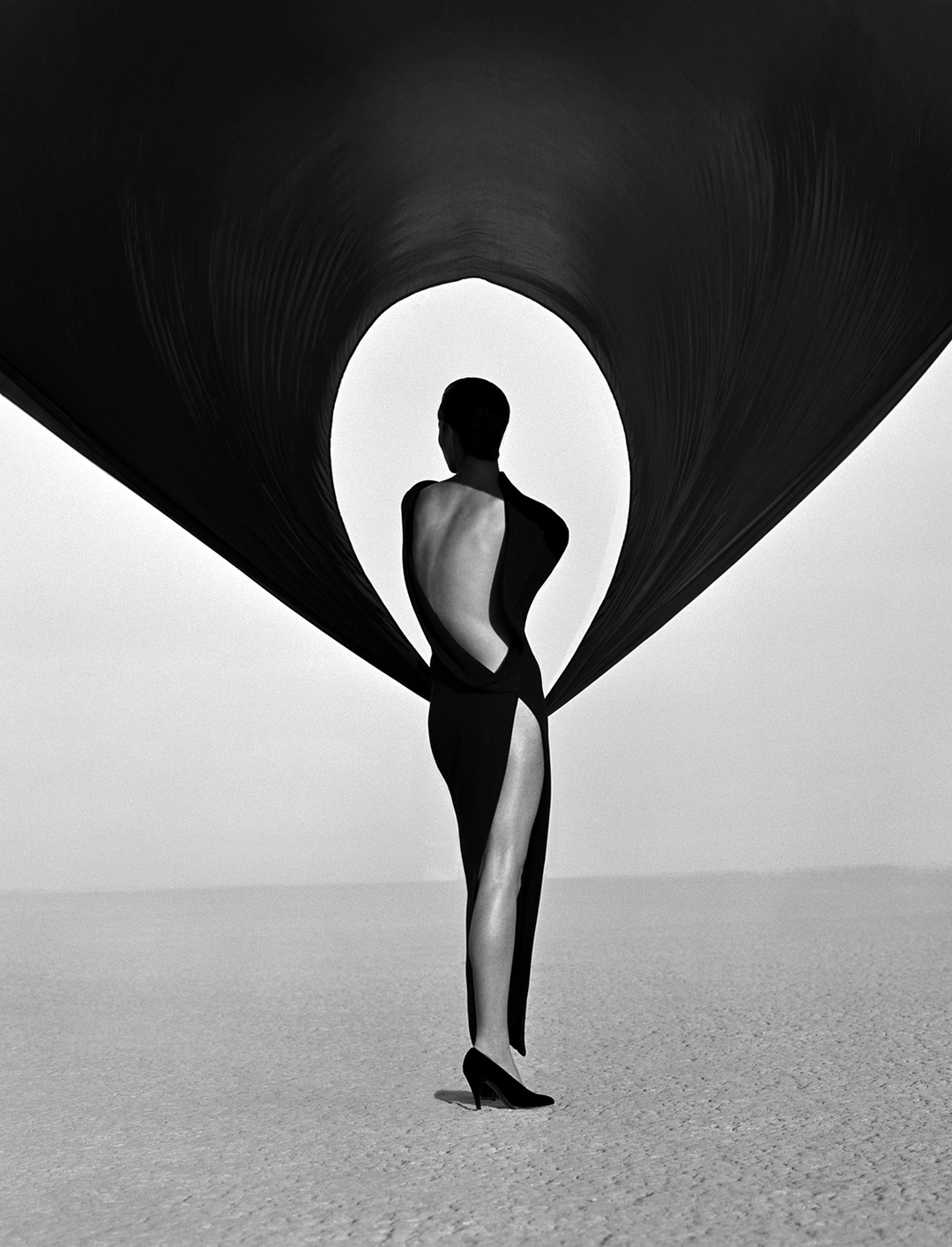Back view of a woman standing in a long black gown whose fabric rises into an enormous oval opening around her upper body in a desert. Herb Ritts “Versace Dress, Back View, El Mirage” 1990 Gelatin silver print Image: 61 x 50.8 cm (24 x 20 in.) Framed: 76.2 x 63.5 cm (30 x 25 in.) El Mirage, California, United States, North America The J. Paul Getty Museum, Los Angeles, Gift of Herb Ritts Foundation © Herb Ritts Foundation