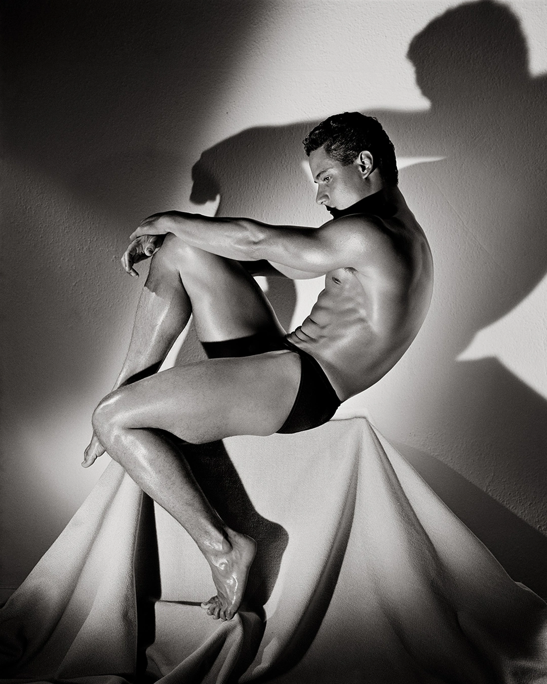 A bare-chested man in dark shorts sits sideways on a draped pedestal, with strong shadows cast onto the wall behind him. Herb Ritts “Greg Louganis, Hollywood” 1987 Gelatin silver print Image: 46 x 38.5 cm (18 1/8 x 15 3/16 in.) Sheet: 50.4 x 40.4 cm (19 13/16 x 15 7/8 in.) Mat: 76 x 63.5 cm (29 15/16 x 25 in.) Hollywood, California, United States, North America The J. Paul Getty Museum, Los Angeles © Herb Ritts Foundation