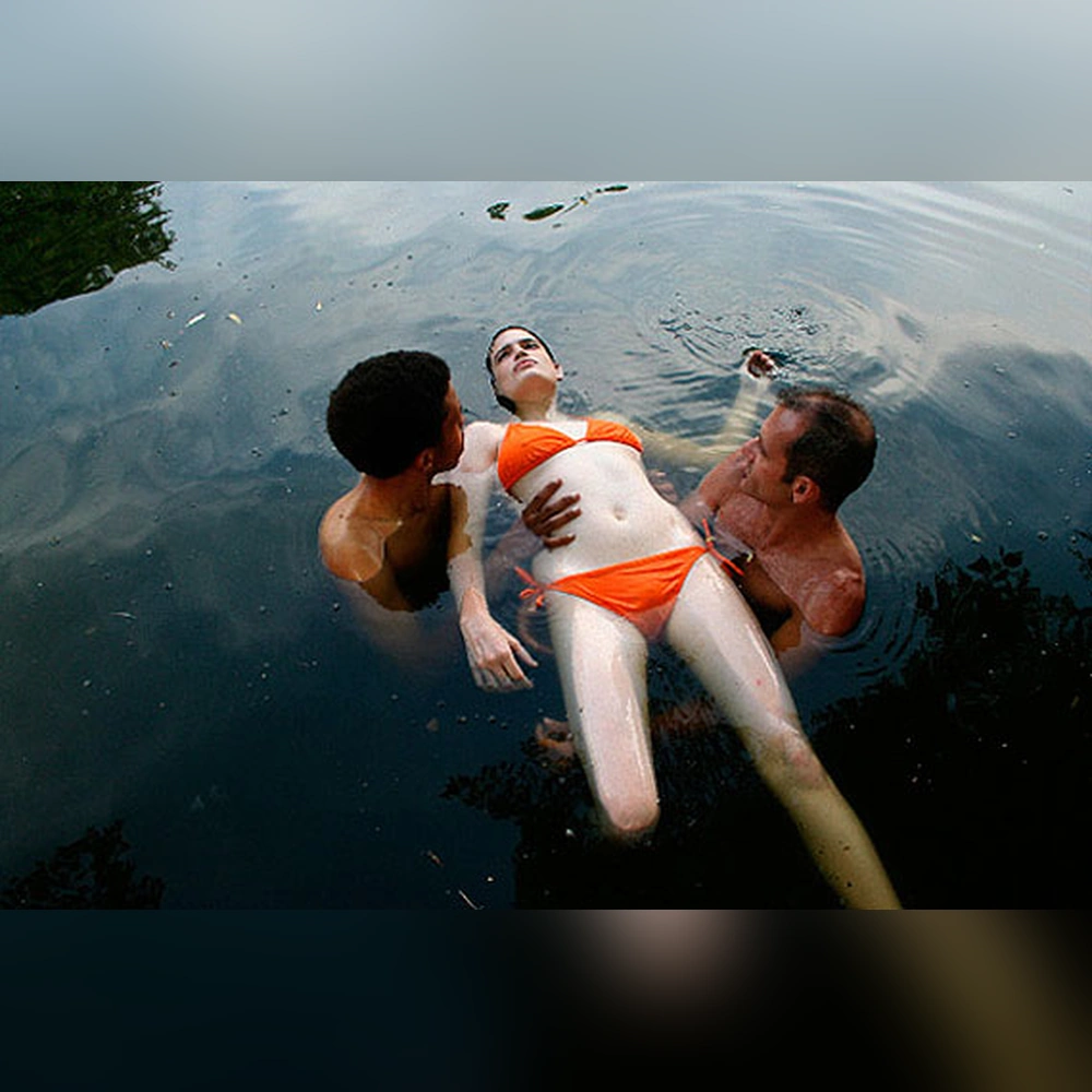 A woman in an orange two-piece swimsuit floats on her back in dark water, supported by two men standing on either side.