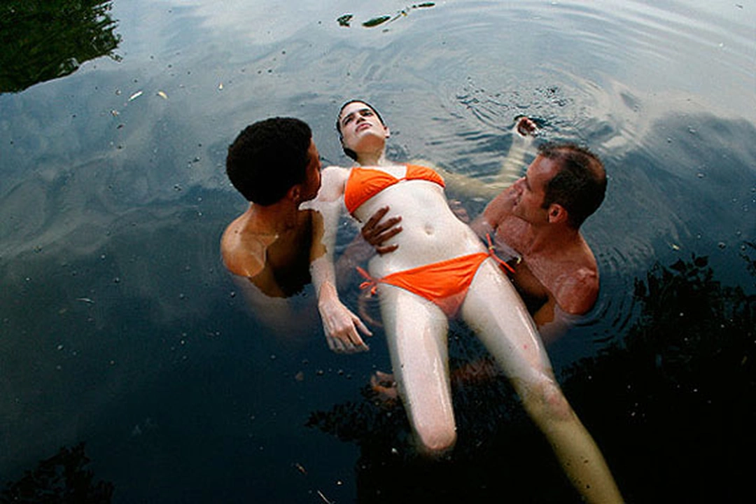 A woman in an orange two-piece swimsuit floats on her back in dark water, supported by two men standing on either side.