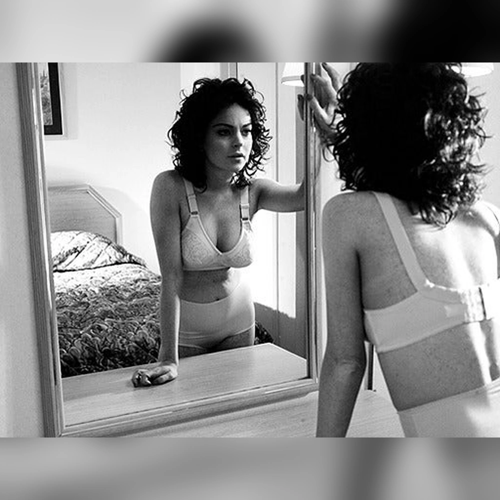 A dark-haired woman in a white bra faces a tabletop mirror, one hand touching the mirror and the other resting beside it, with a bed behind her.