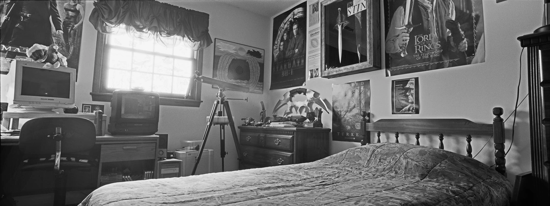 Black-and-white panoramic view of a bedroom with a bed in the foreground, a computer desk by the window and posters covering the walls.