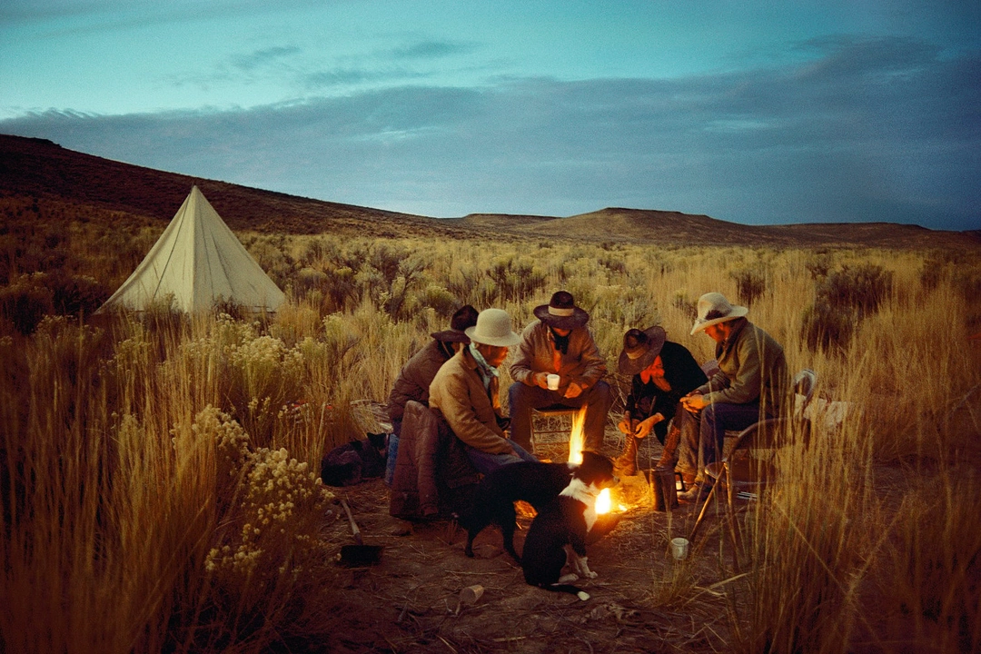 Cowboys gather around a campfire beside a tent in tall grass beneath a blue dawn sky. William Albert Allard, “Cow Camp at Dawn”