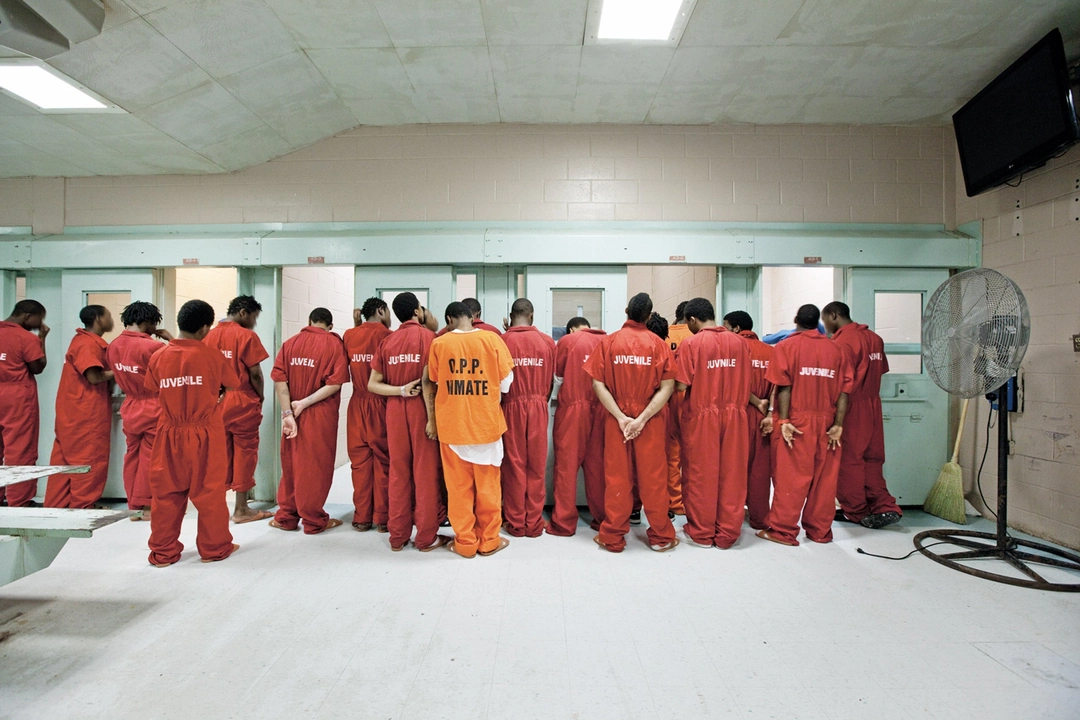 Young people in red JUVENILE uniforms stand with their backs to the camera in a detention facility; an orange uniform stands among them and a large fan is at the right. Richard Ross, “Red Suits”