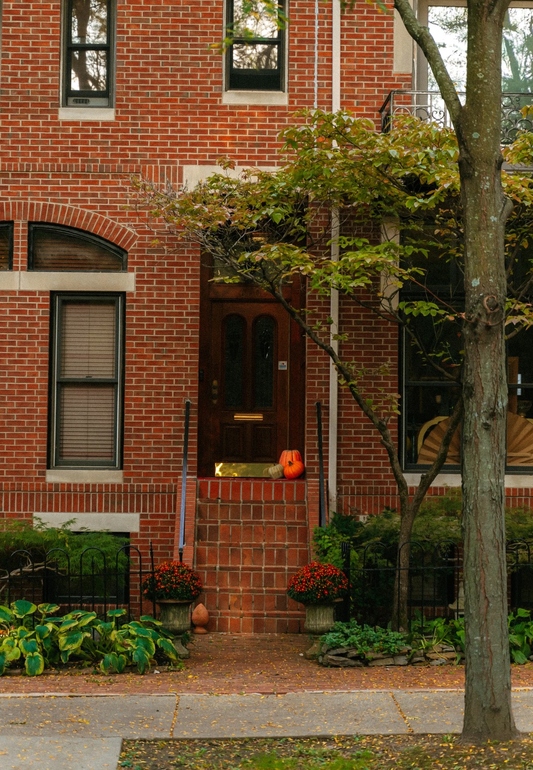 Brick house with pumpkins and red chrysanthemums on the porch, autumn tree and leaf-covered sidewalk.