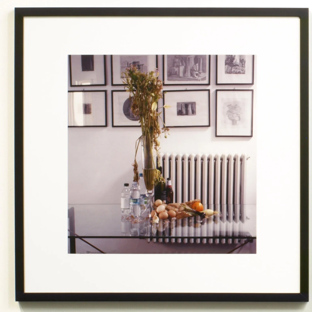 Glass dining table with food and bottles under a tall floral arrangement, in front of a radiator and framed etchings.