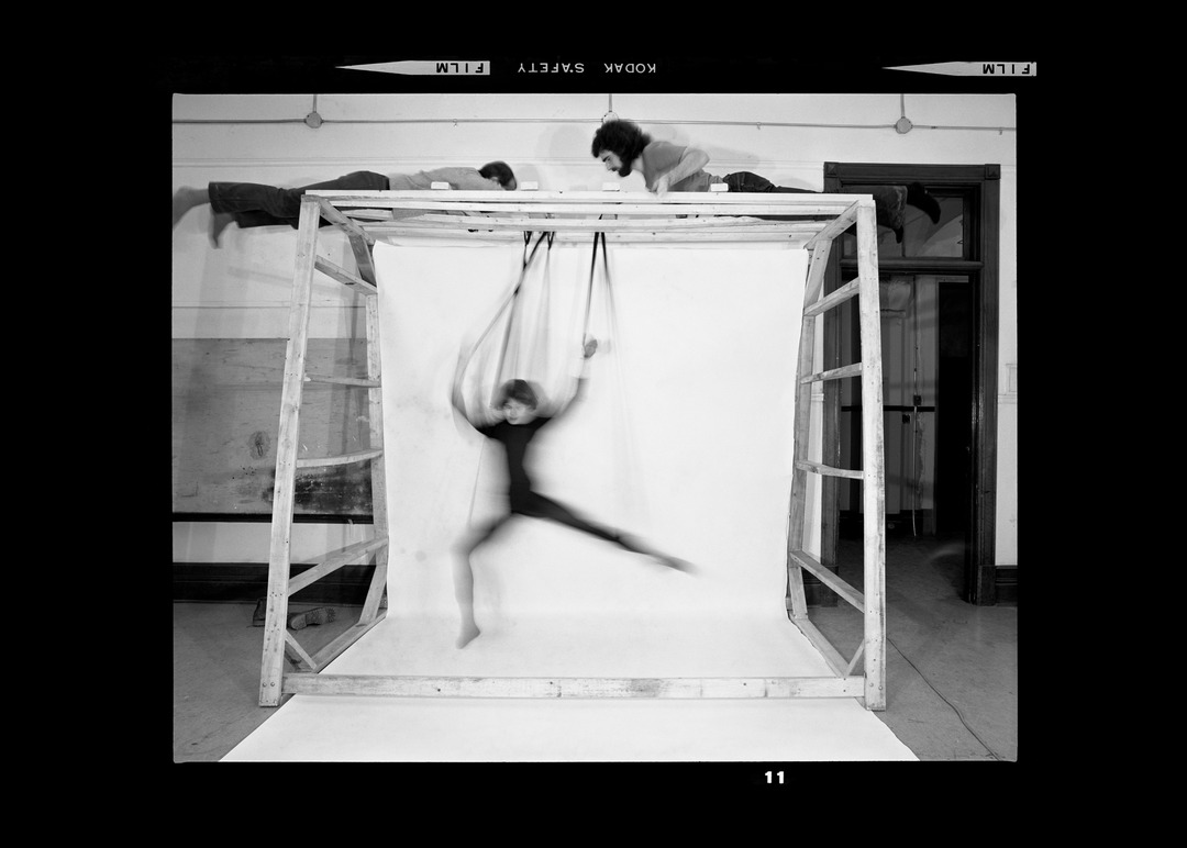 Black-and-white negative scan of a performer suspended mid-air inside a wooden frame structure.