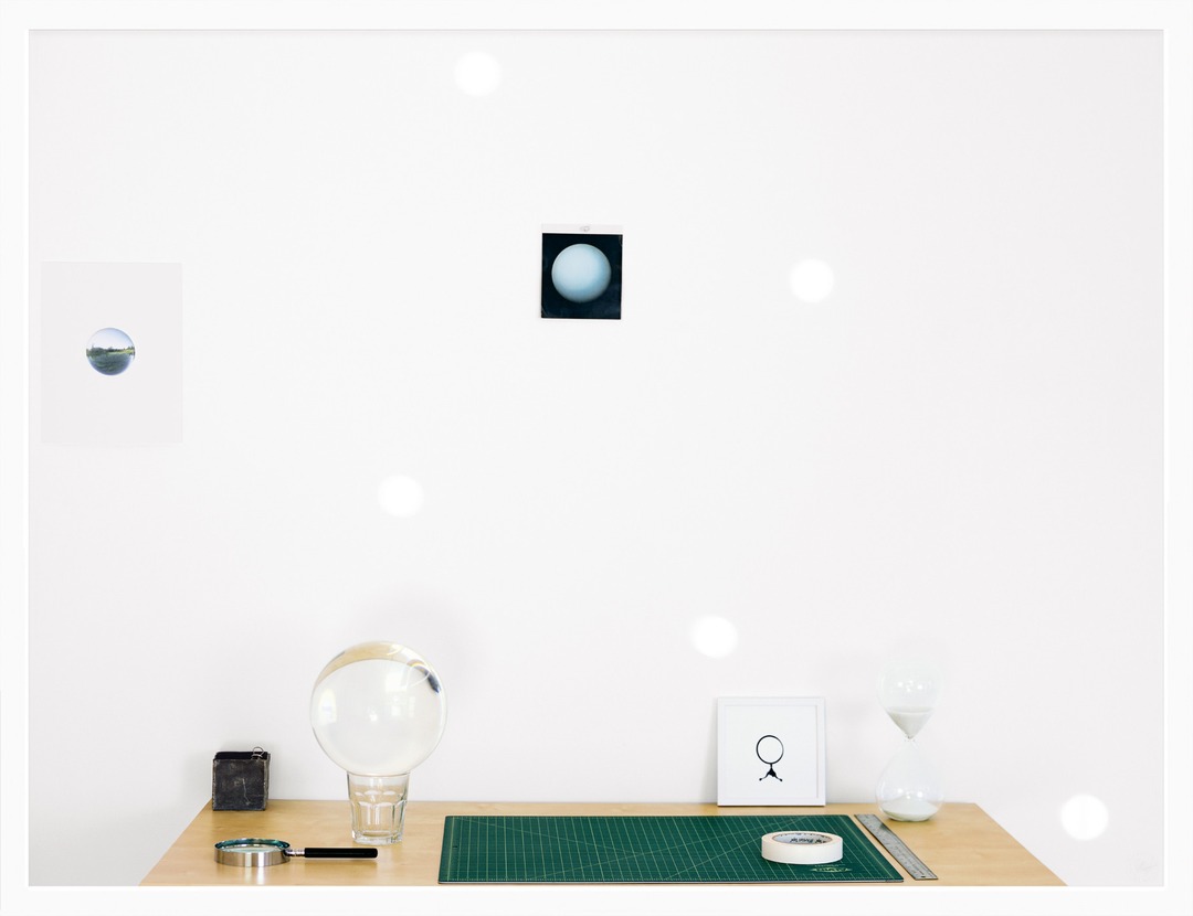 A studio table with a large glass sphere, cutting mat, hourglass and small framed prints on a white wall.