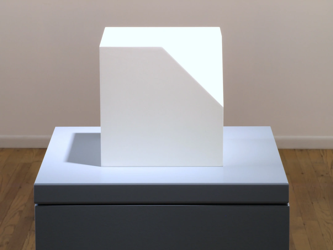 White cube sculpture with a triangular corner removed stands on a gray rectangular pedestal. Sol LeWitt, Cube Without a Corner, 2005, Syntactic Polyurethane slabs with lacquer, 6 x 6 x 6 inches.