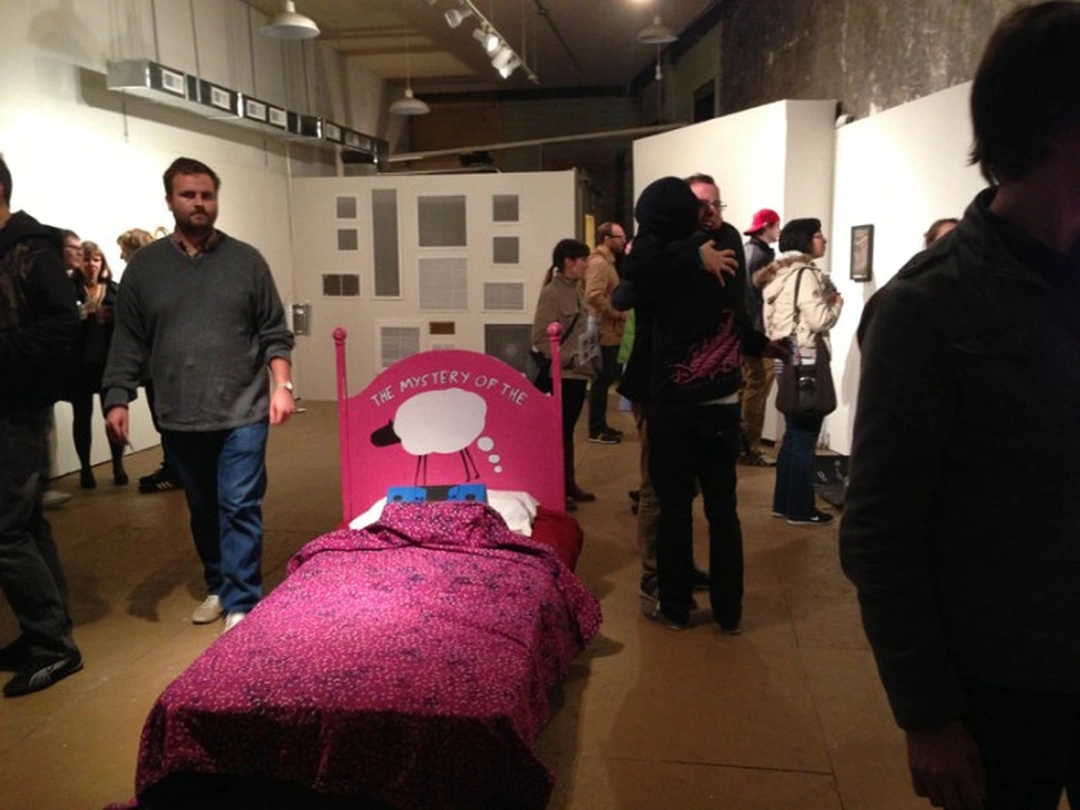 A pink bed with patterned bedding and a speech-bubble headboard stands among visitors in an industrial gallery, with rectangular grilles arranged on the wall behind it.
