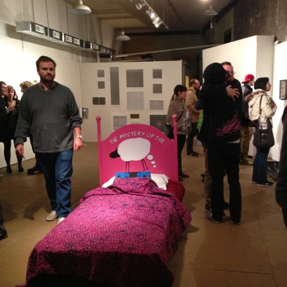 Pink bed with patterned bedding and a speech-bubble headboard surrounded by visitors at a gallery closing exhibition.