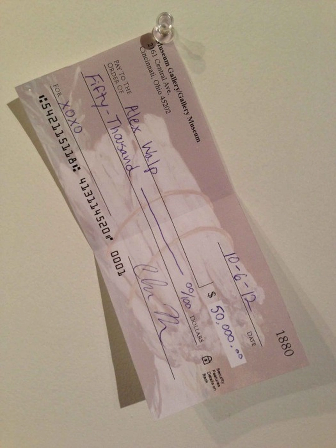 A pale pink personal check hangs diagonally against a beige wall, suspended from a small pin near its upper edge.