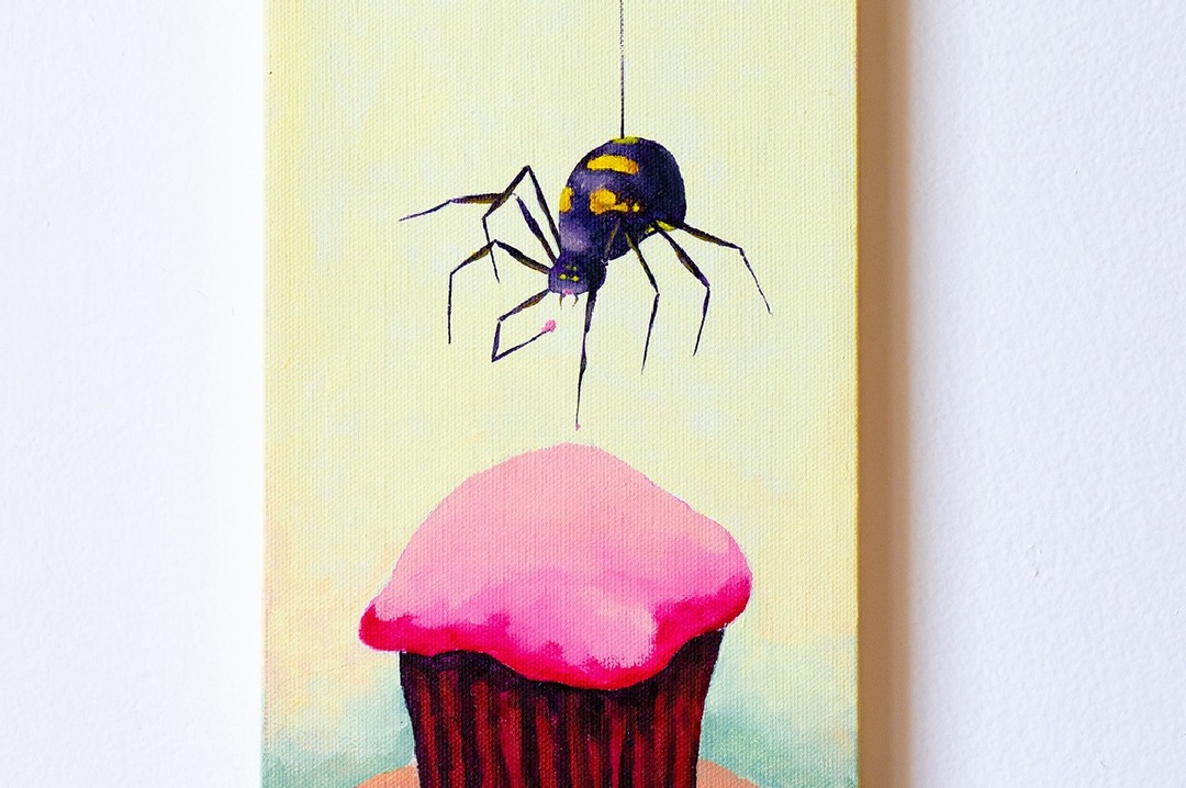 Photograph of a painting of a black-and-yellow spider dangling from a thread above a pink-frosted cupcake, against a pale yellow wall.