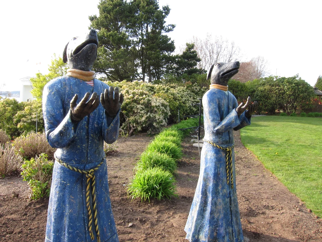 Two sculptures of dog-headed figures in blue robes, hands raised, standing in the gardens of Peace Arch Park on the Canada-United States border.