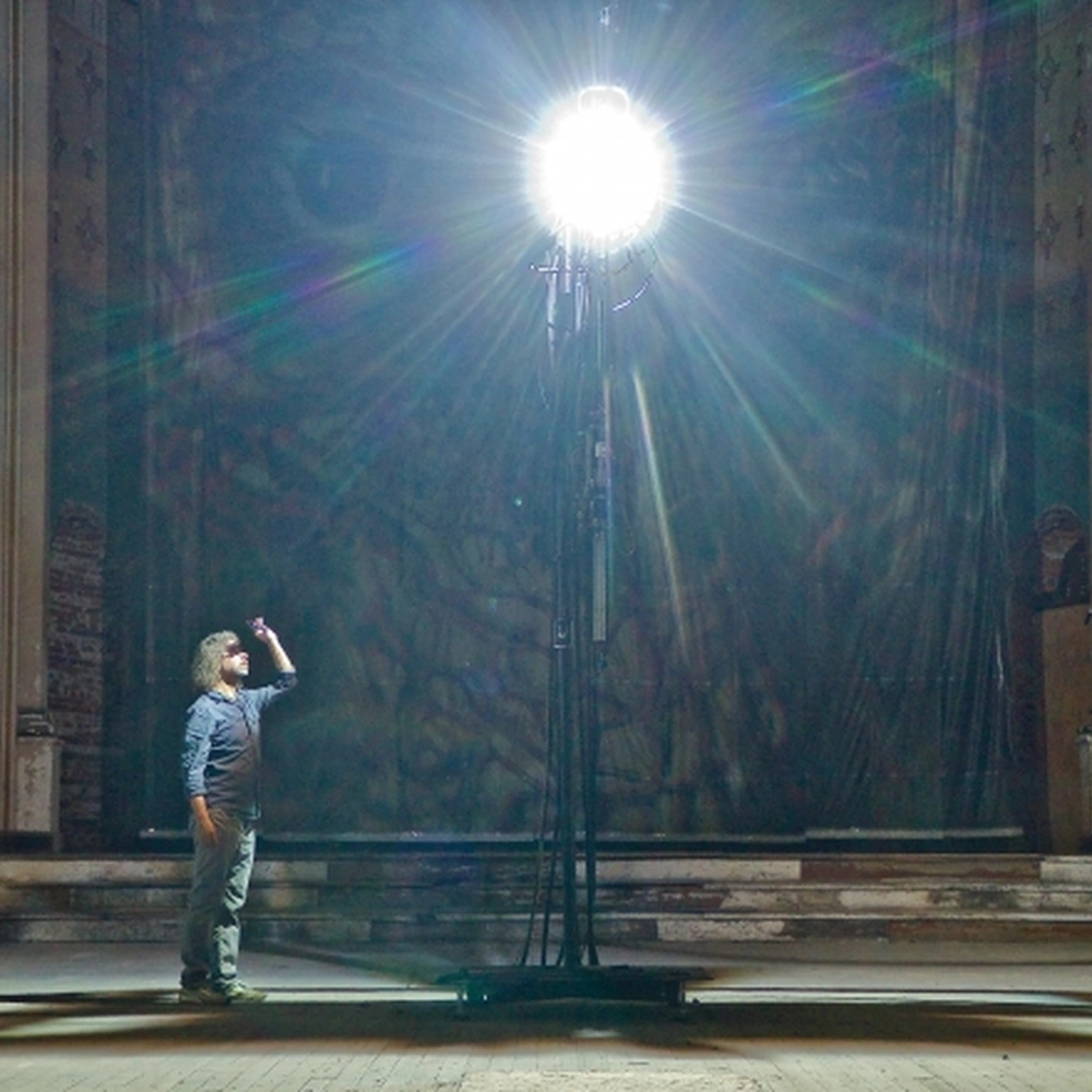 A visitor stands beneath a brilliant carbon arc lamp in front of a monumental photographic work in the apse of a worn church interior.