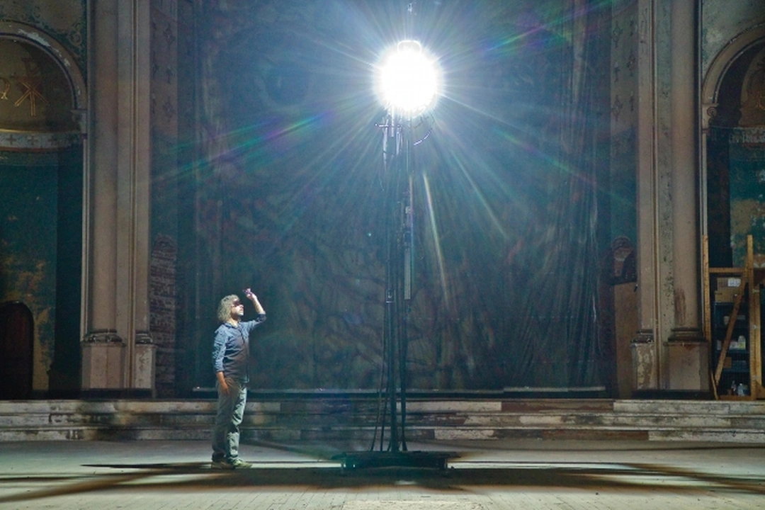 A visitor stands beneath a brilliant carbon arc lamp in front of a monumental photographic work in the apse of a worn church interior.