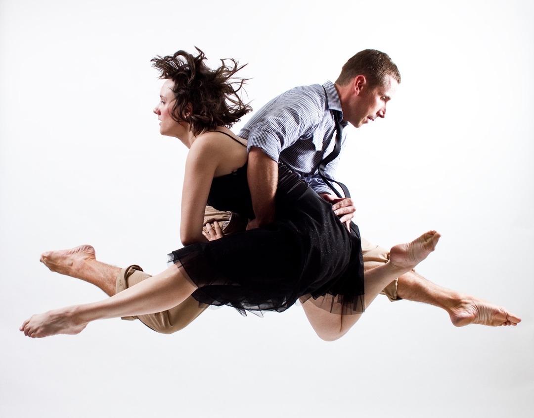 Dance duet in the air: female dancer in a black dress held aloft by a male dancer, white background.