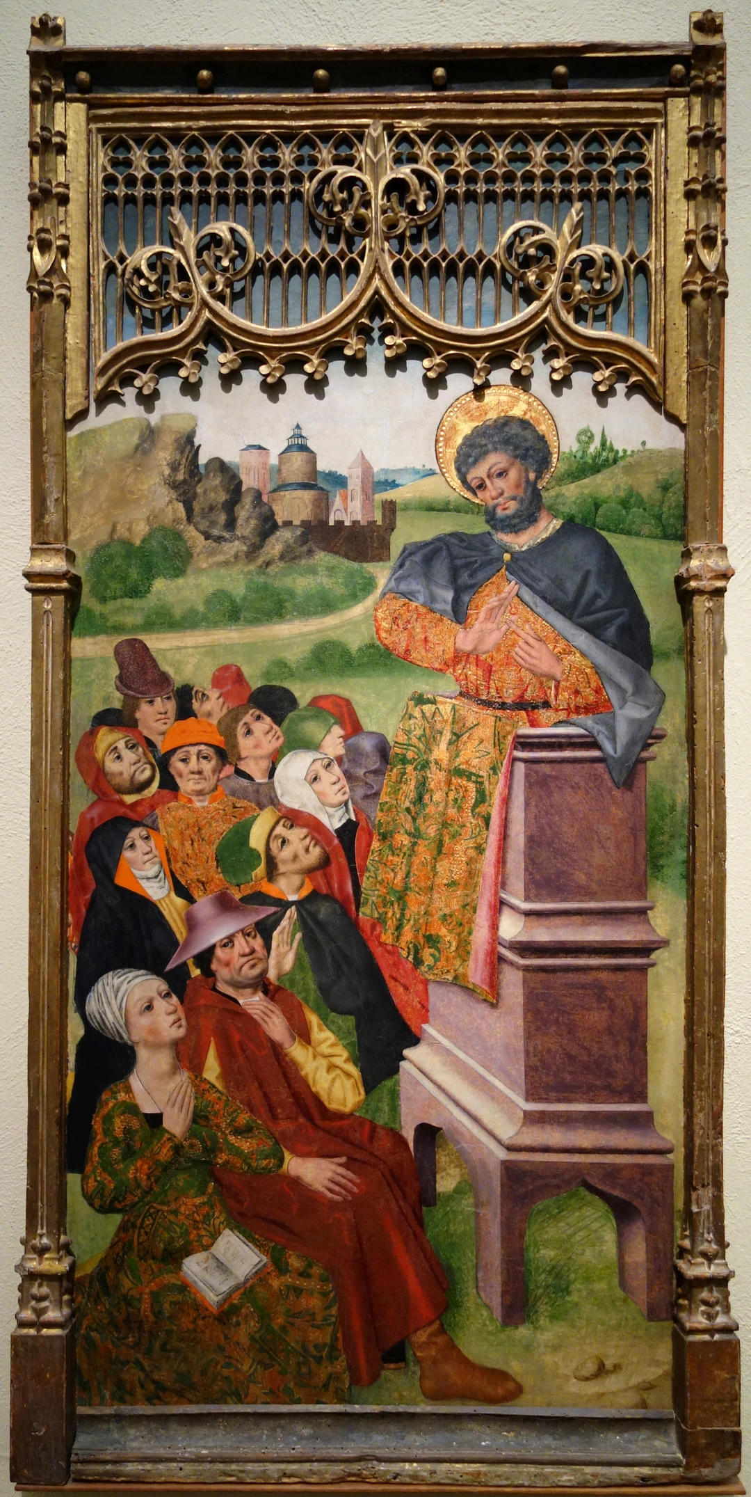 Fifteenth-century Spanish panel painting in a gilded Gothic frame: a haloed saint preaches from a pulpit to a crowd of men and women in colorful hats and robes, with a hilltop castle in the landscape behind.