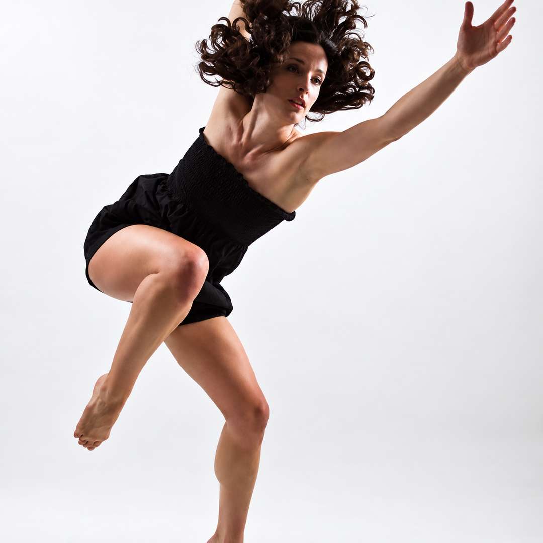 Dancer mid-leap against a white background, black leotard, arm raised, curly hair in motion.