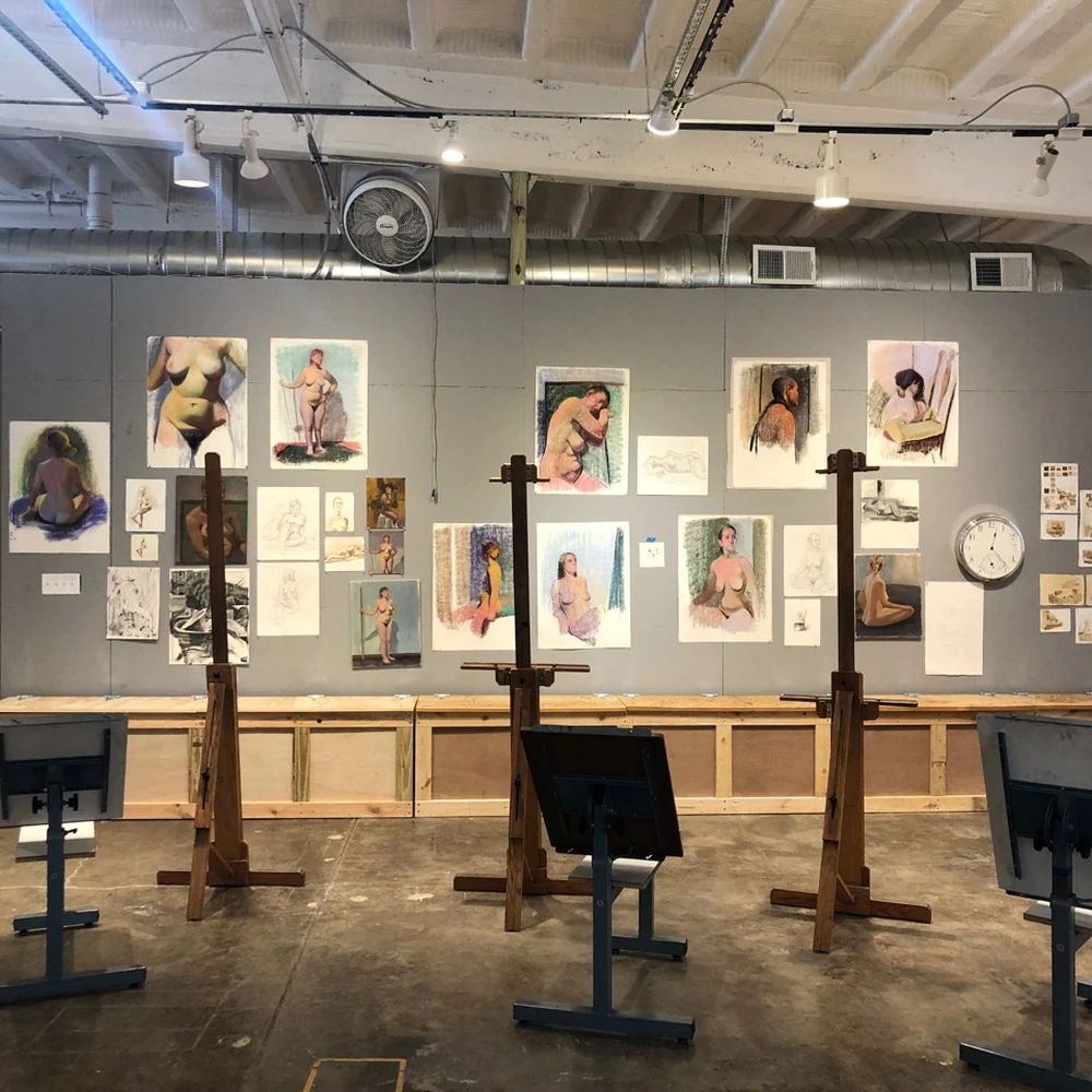 A drawing studio with wooden easels and figure studies pinned to a gray wall.