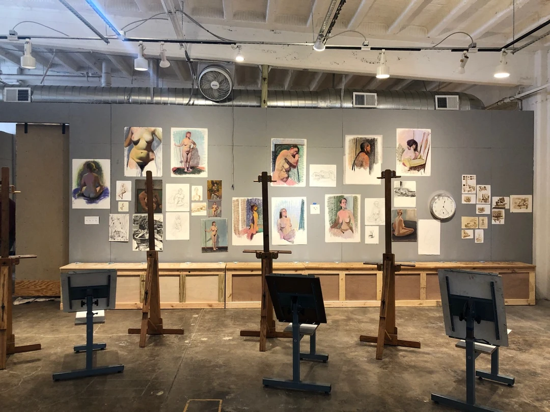 A drawing studio with wooden easels and figure studies pinned to a gray wall.