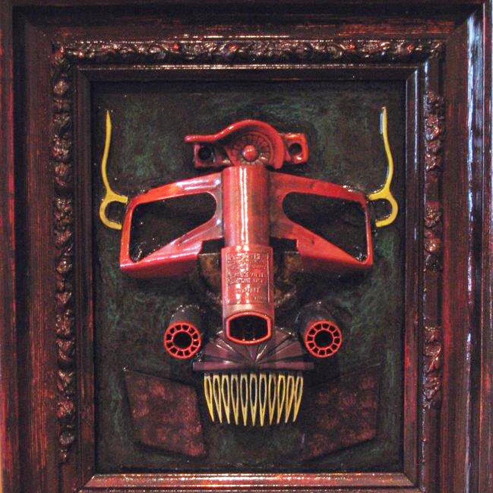 Framed assemblage by Jeff Casto, Wasteland Warrior: a red mask-like face built from found objects — a tall central column flanked by curved side pieces, round red-rimmed eyes and a grinning row of yellow teeth — set on a dark textured ground inside a carved dark wood frame.