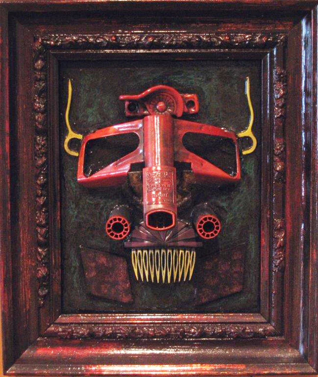 Framed assemblage by Jeff Casto, Wasteland Warrior: a red mask-like face built from found objects — a tall central column flanked by curved side pieces, round red-rimmed eyes and a grinning row of yellow teeth — set on a dark textured ground inside a carved dark wood frame.
